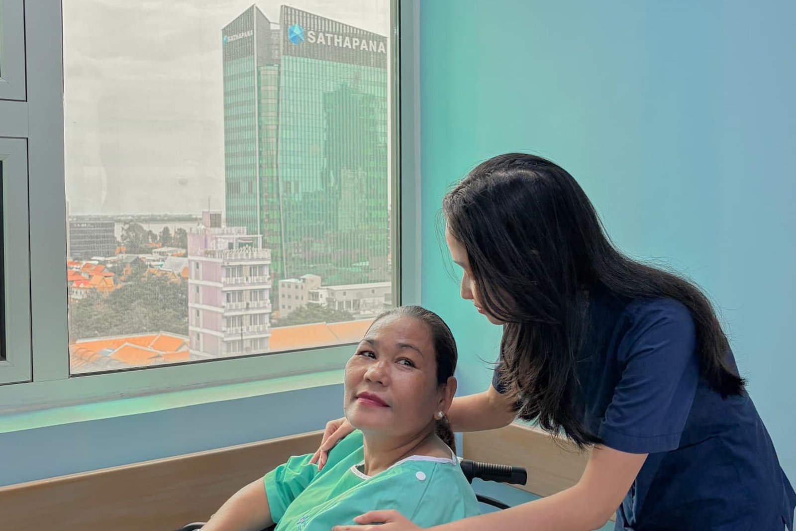 A Metta Lotus caregiver in navy uniform standing beside a woman seated in a wheelchair by a sunlit window