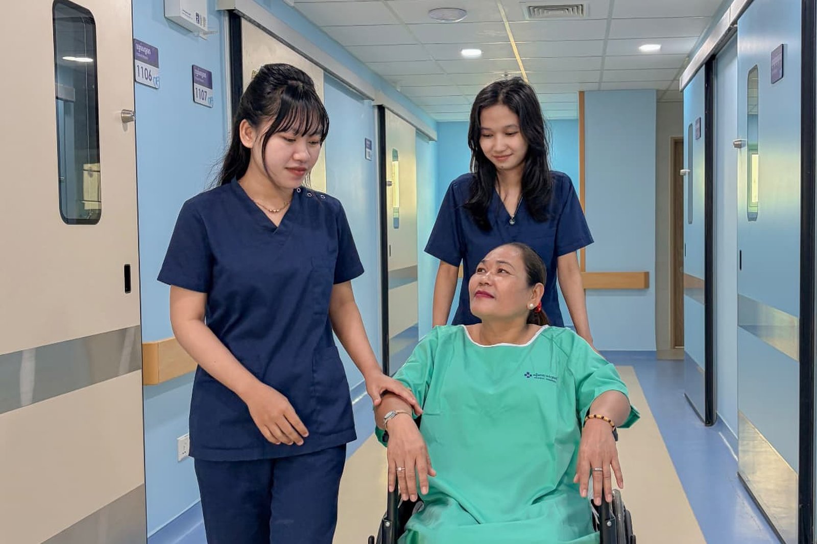 Two Metta Lotus caregivers in navy uniform accompanying a woman in a wheelchair along a bright corridor