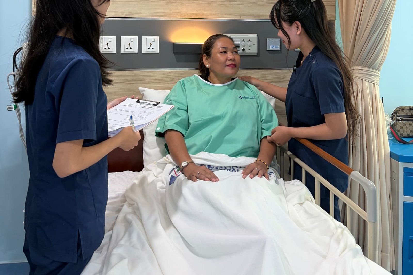 Two Metta Lotus caregivers in navy uniform at the bedside of a resting patient, one noting care details on a chart