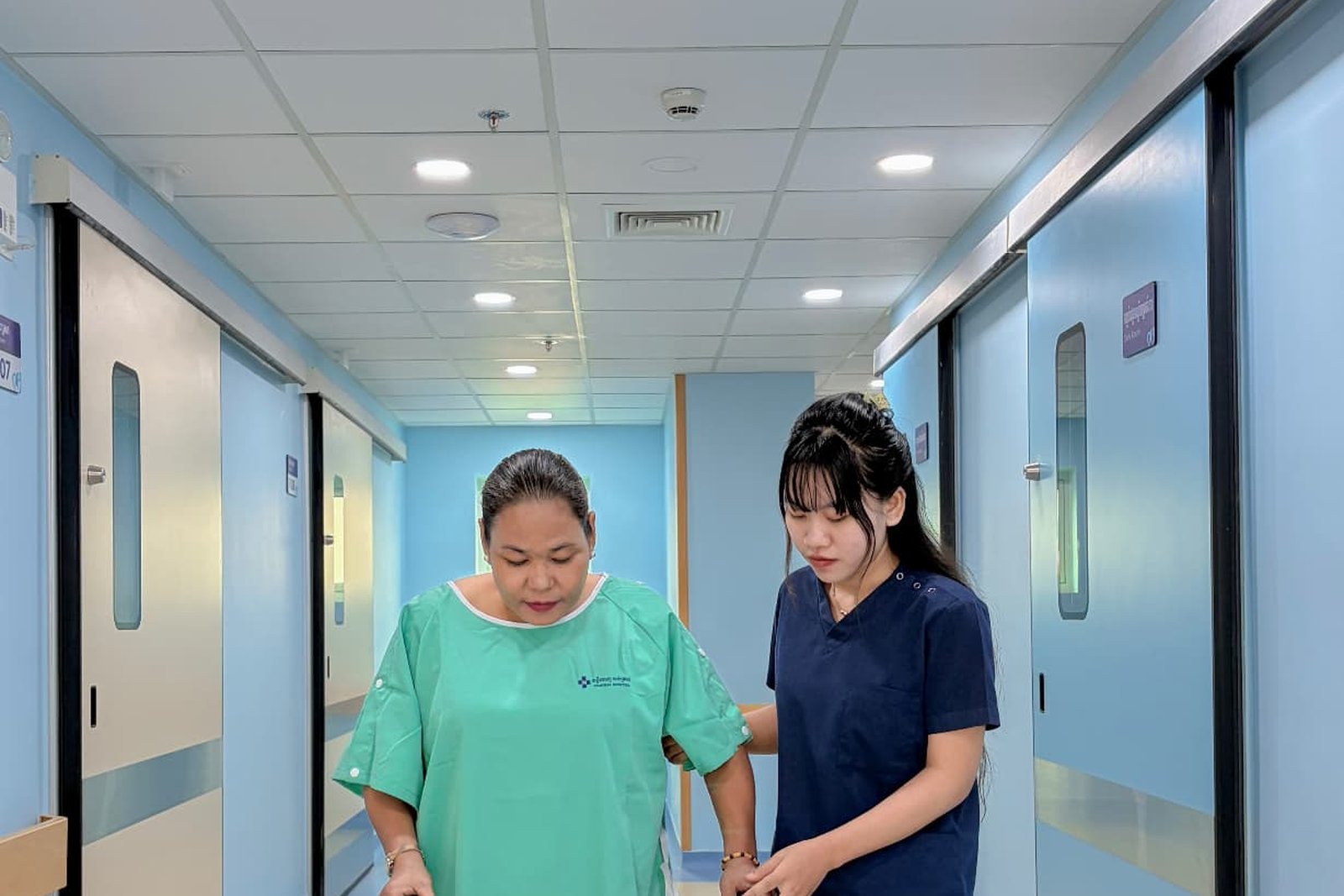 A Metta Lotus caregiver in navy uniform steadying a woman as she walks slowly along a corridor